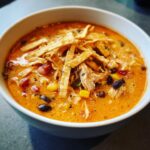 A close-up of a bowl of creamy, orange-hued tortilla soup recipe, topped with shredded chicken, black beans, corn, and crispy tortilla strips.