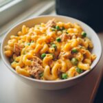 Close-up of creamy tuna pasta mixed with green peas and melted cheddar cheese in a white bowl.