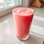 A tall glass filled with a vibrant, creamy watermelon smoothie sitting on a white marble counter near a window.