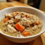 A close-up of creamy wild rice soup featuring chunks of chicken and bright orange carrots in a white bowl.