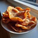 A close-up of crispy, golden-brown buñuelos generously dusted with sugar, piled high in a white bowl near a window.