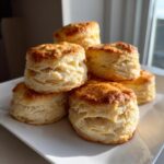 A stack of golden-brown, flaky buttermilk biscuits piled on a white plate near a window.