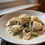 A close-up of a bowl of fluffy chicken and dumplings served in a creamy sauce with bright green peas.
