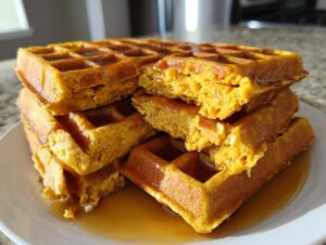 A close-up stack of fluffy, golden-orange pumpkin waffles drizzled generously with maple syrup.
