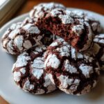 A close-up of several fudgy chocolate crinkle cookies dusted heavily with white powdered sugar on a white plate.