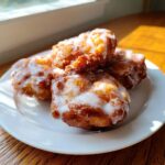 A stack of four golden brown, glazed apple fritter pastries sitting on a white plate near a window.
