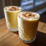 Two tall, textured glasses filled with creamy homemade eggnog, topped with a sprinkle of cinnamon.