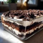 Close-up of a layered dirt cake in a glass dish, showing alternating layers of chocolate cake, pudding, and whipped cream, topped with crushed Oreos.