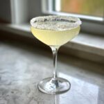 A bright yellow lemon drop martini served in a coupe glass with a thick, sugared rim, sitting on a marble counter.