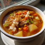 A close-up of a bowl of Maryland crab soup, featuring large chunks of crab meat, potatoes, carrots, and corn, topped with paprika.
