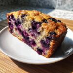 A thick slice of moist blueberry banana bread with a golden, sugary crust, served on a white plate.