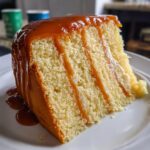 Close-up of a moist slice of vanilla caramel cake layered with cream and generously topped with dripping caramel sauce.