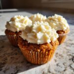 Close-up of four moist carrot cake muffins topped with thick swirls of white cream cheese frosting.