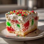 A moist slice of christmas poke cake showing red and green jello pockets, topped with whipped cream and festive sprinkles.