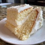 Close-up of a moist slice of layered vanilla cake recipe covered in thick white buttercream frosting.