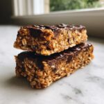 Two stacked no bake granola bars made with oats and topped with melted dark chocolate, resting on a marble surface.