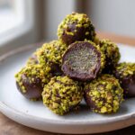 A pile of rich, dark chocolate dubai chocolate balls coated in bright green chopped pistachios, with one cut in half showing the textured filling.