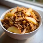 A white bowl filled with tender shredded pork and sauerkraut, garnished with fresh apple slices.