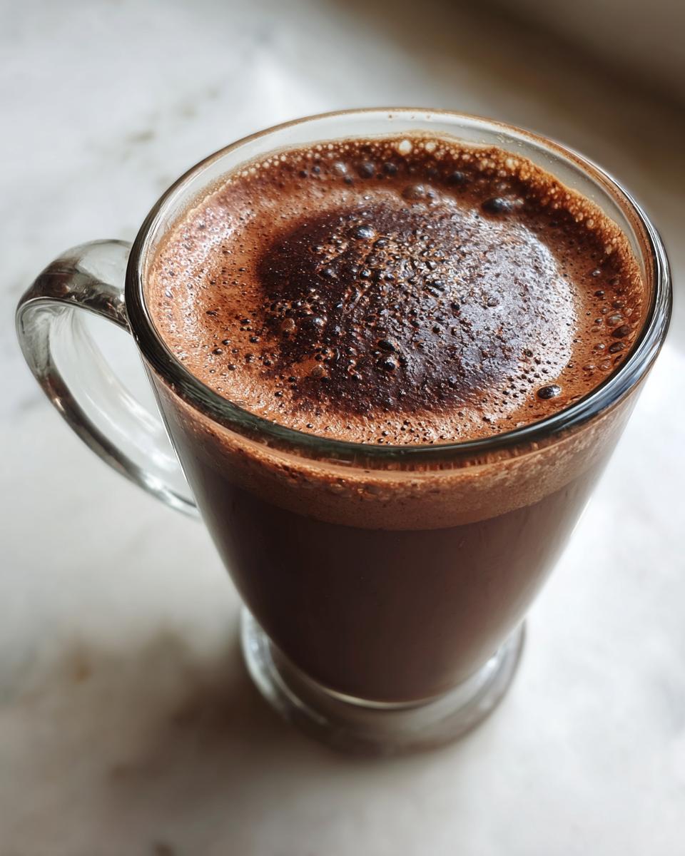 Close-up of a mug filled with rich, dark hot cocoa mix topped with thick, dark brown foam.