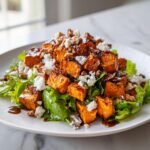 Close-up of a vibrant sweet potato salad featuring roasted orange cubes, crumbled white cheese, pecans, and dark glaze over greens.