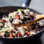 A wooden spoon lifts a generous serving of flavorful black beans and rice mixed with sausage pieces from a skillet.