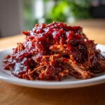 A close-up of tender, shredded Slow cooker pulled pork piled high and generously coated in thick barbecue sauce on a white plate.