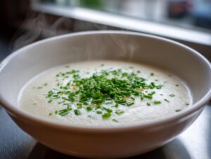 Close-up of a steaming white bowl filled with creamy cauliflower soup, topped generously with fresh green chives.