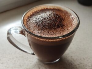 Close-up of a clear glass mug filled with rich, dark hot cocoa topped with foam, made from hot cocoa mix.