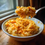 A fork lifts a portion of incredibly cheesy rice from a white bowl, showing long cheese pulls of melted cheddar cheese.
