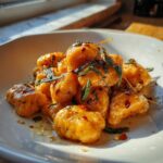 A close-up of glossy, orange sweet potato gnocchi coated in brown butter sauce, topped with crispy sage leaves and red pepper flakes.