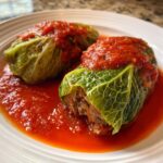 Close-up of two savory cabbage rolls stuffed with meat, covered in bright red tomato sauce, served on a white plate.