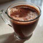 Close-up of a rich, dark hot cocoa mix beverage in a clear glass mug, topped with foam and dusted cocoa powder.