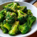 Close-up of bright green steamed broccoli florets tossed with seasoning in a white bowl.