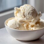 Three scoops of creamy, light-colored protein ice cream served in a white bowl, highlighted by sunlight.