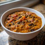 A close-up of a white bowl filled with thick, orange vegan pumpkin chili featuring black beans and diced tomatoes.