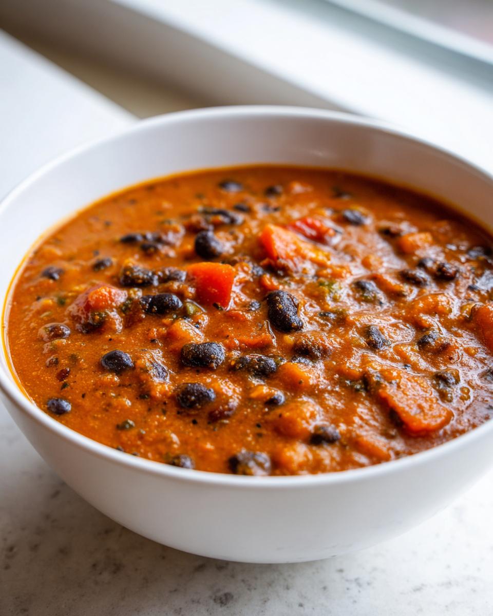 Close-up of a white bowl filled with thick, orange-red vegan pumpkin chili featuring black beans and chunks of sweet potato.