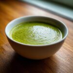 A close-up of vibrant green asparagus soup served in a white bowl on a wooden surface.