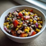 Close-up of colorful cowboy caviar featuring black beans, corn, tomatoes, and red onion in a white bowl.