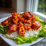 Close-up of vibrant, saucy harissa chicken pieces served over white rice and surrounded by fresh green lettuce leaves.