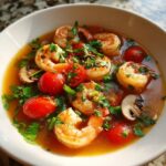 A close-up of a bowl of tom yum soup featuring plump shrimp, cherry tomatoes, mushrooms, and cilantro.