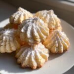 A stack of delicate, piped whipped shortbread cookies dusted heavily with powdered sugar on a white plate.