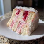 A moist slice of white chocolate raspberry cake showing layers of white cake, bright red raspberry filling, and white frosting.