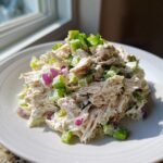 A mound of creamy, shredded chicken salad mixed with diced celery, red onion, and topped with green onions.
