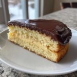 A close-up of a thick slice of Boston Cream Pie featuring yellow cake, vanilla pastry cream, and dark chocolate ganache.