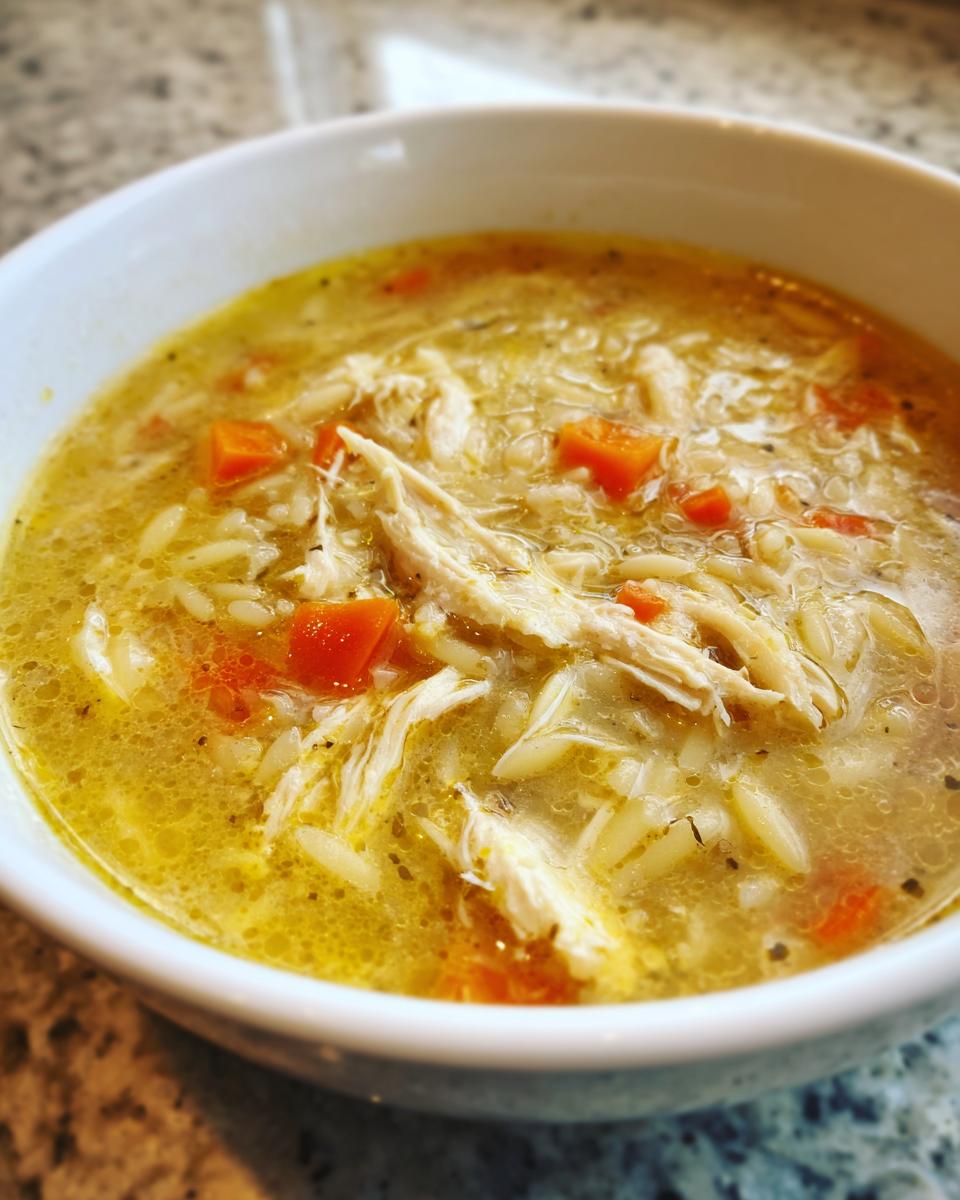 Close-up of a white bowl filled with rich, yellow chicken orzo soup, featuring shredded chicken, carrots, and herbs.