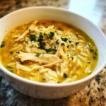 Close-up of a white bowl filled with rich, yellow broth chicken orzo soup, topped with shredded chicken and fresh parsley.