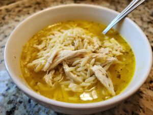 Close-up of a white bowl filled with flavorful chicken orzo soup, featuring shredded chicken and orzo pasta in a rich yellow broth.