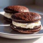 Two delicious chocolate whoopie pies filled with thick white cream frosting, sitting on a white plate.