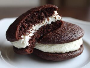 A close-up of two chocolate whoopie pies filled with thick white cream, one piece broken open.