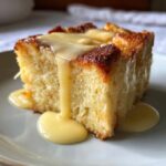 A close-up of a square slice of golden brown bread pudding being generously drizzled with vanilla custard sauce.